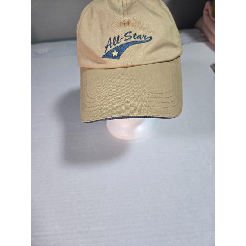 May include: A tan baseball cap featuring the embroidered text All-Star in dark blue script. The design includes a dark blue swoosh shape underneath the text with a small yellow star centered within it. The cap has a curved brim and stitching.