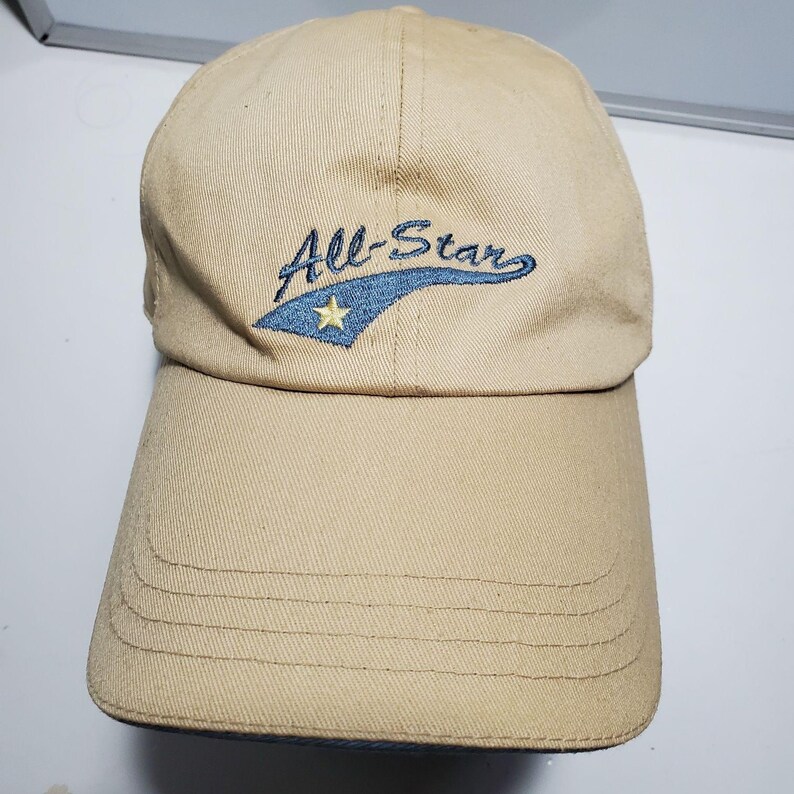 May include: A tan baseball cap featuring an embroidered design on the front panel. The embroidery includes the text All-Star in dark blue script, positioned above a blue swoosh shape that contains a small, yellow five-pointed star icon.