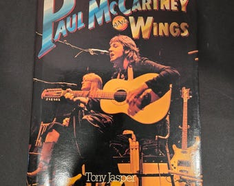 May include: A book cover featuring a photo of Paul McCartney and Wings performing live on stage. The cover is black with white text that reads "Paul McCartney and Wings" and "Tony Jasper". The text "over 100 illustrations in color and black and white" is also visible.
