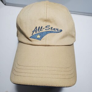 Könnte beinhalten: Eine beigefarbene Baseballkappe mit einem gestickten Motiv auf der Vorderseite. Die Stickerei zeigt den Schriftzug All-Star in dunkelblauer Schreibschrift über einem blauen geschwungenen Element, das einen kleinen gelben fünfzackigen Stern enthält.