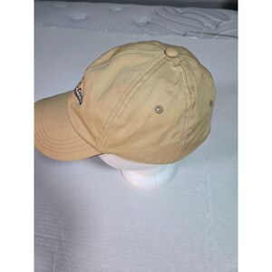 May include: A tan baseball cap featuring a curved brim and a button at the crown. The side of the cap displays a partial blue embroidered logo with the visible text "Star". The cap is constructed with stitched panels and includes small metal eyelets.
