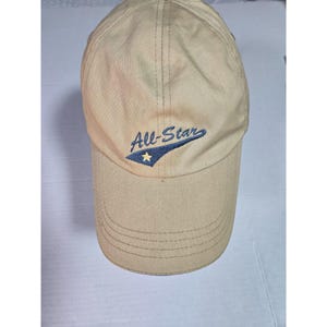 May include: A tan baseball cap featuring the embroidered text All-Star in blue script. The design includes a blue pennant-shaped graphic underlined with a small yellow star. The cap has a structured crown and a curved brim with visible stitching.