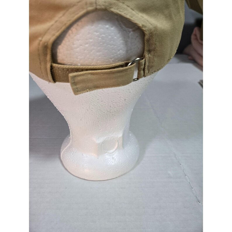 May include: A tan baseball cap featuring an adjustable strap closure at the back. The cap is constructed from a solid-colored fabric and includes a metal buckle mechanism for securing the fit. The design includes a rear opening above the strap.