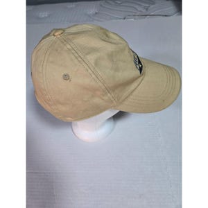 May include: A tan baseball cap featuring a structured crown with a button top, stitched eyelets, and a curved brim. A dark blue patch with white text and a small star graphic is positioned on the front side of the cap, which is displayed on a stand.