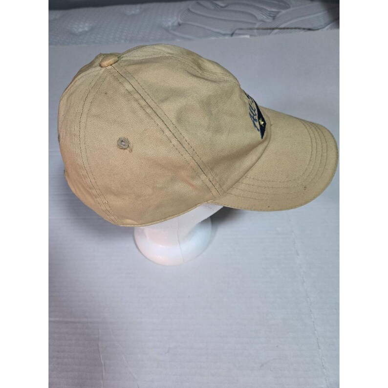 May include: A tan baseball cap featuring a structured crown with a button top, stitched eyelets, and a curved brim. A dark blue patch with white text and a small star graphic is positioned on the front side of the cap, which is displayed on a stand.