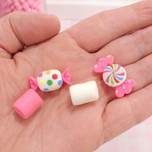 May include: A hand holds miniature candy-themed polymer clay charms. The charms include a white candy with colorful polka dots, a pink cylinder, a white marshmallow, and a pink candy with a swirled rainbow design. A glass jar filled with pink candies is in the background.