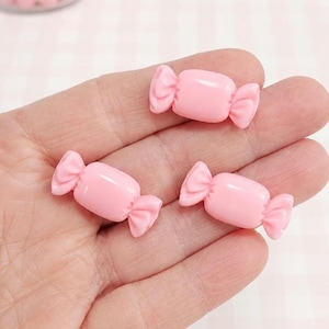 May include: Three pink candy-shaped embellishments with a wrapped design are held in a hand. The candies are a solid, light pink color and have a bow-like detail on each end. A jar of pink beads is in the background.