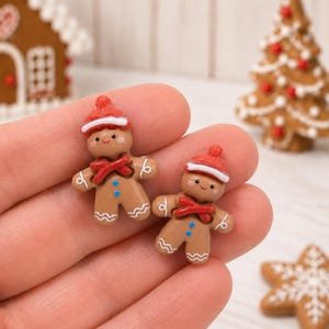 May include: Two small gingerbread person figurines made of brown material, each wearing a red hat with a white brim and a red scarf. They feature white icing details on their arms and legs, two blue dots on their torsos, and painted faces.