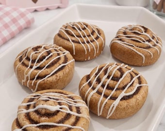 May include: Five round cinnamon rolls arranged on a white rectangular tray. Each golden-brown pastry features a spiral pattern filled with dark brown cinnamon and is drizzled with white icing. A pink sign in the background reads Cinnamon Rolls.
