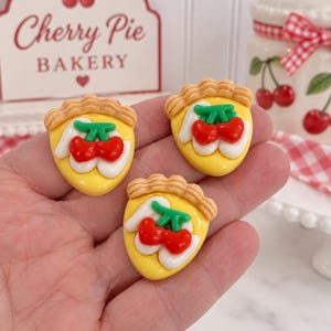 May include: Three small, triangular-shaped cherry pie charms, each featuring a yellow crust, white filling, and two bright red cherries with green stems on top. The charms have a textured, scalloped edge along the top crust section.