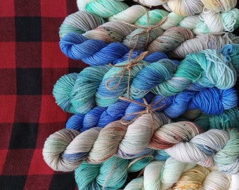May include: A stack of colorful yarn skeins in shades of blue, green, white, and brown. The yarn is arranged in a pyramid shape and tied together with twine.