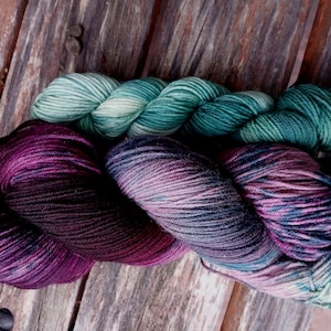 May include: Close-up of three skeins of yarn in shades of teal, purple, and pink. The yarn is arranged on a weathered wooden surface. The yarn appears soft and is likely used for knitting or crocheting projects.