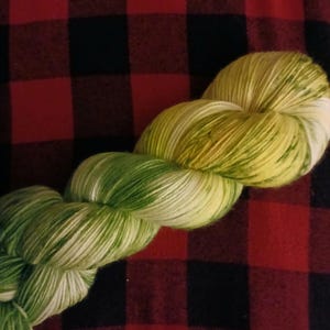 May include: A twisted skein of yarn featuring a variegated color palette of bright lime green, soft yellow, and crisp white. The fibers are tightly wound into a long, braided-style hank, showcasing a blend of light and dark green tones.