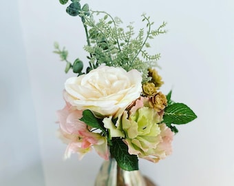 May include: A bouquet of artificial flowers in a clear glass vase. The bouquet includes a large white rose, pink flowers, and green foliage.