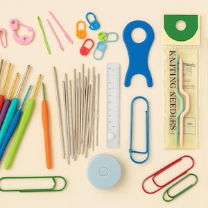 Pode incluir: Uma cole&ccedil;&atilde;o de ferramentas coloridas de croch&ecirc; e tric&ocirc;, incluindo agulhas de croch&ecirc; com cabos coloridos, agulhas de metal, marcadores de ponto, clipes de papel e uma fita m&eacute;trica. As ferramentas est&atilde;o dispostas sobre uma superf&iacute;cie bege clara.