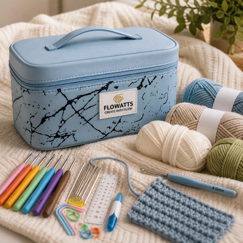 Pode incluir: Uma caixa de armazenamento artesanal azul claro com uma al&ccedil;a, design preto semelhante a m&aacute;rmore e o texto "FLOWATTS CREATE WITH FLOW". Agulhas de croch&ecirc; sortidas, fios e uma amostra de croch&ecirc; s&atilde;o dispostos em uma superf&iacute;cie creme.