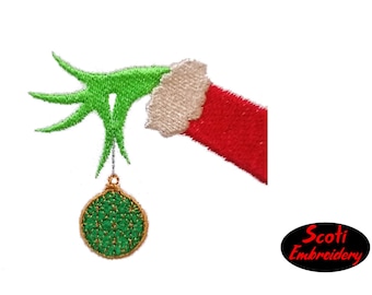 Op de afbeelding: Groene hand met een rode mouw die een groene en gouden kerstbal vasthoudt. De tekst "Scoti Embroidery" staat in een zwarte ovaal.