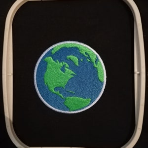 May include: Embroidered patch showing the Earth, with green continents and blue oceans. The circular patch has a white border and is set against a black background within an embroidery hoop. Suitable for customising jackets, bags, or other fabric items.
