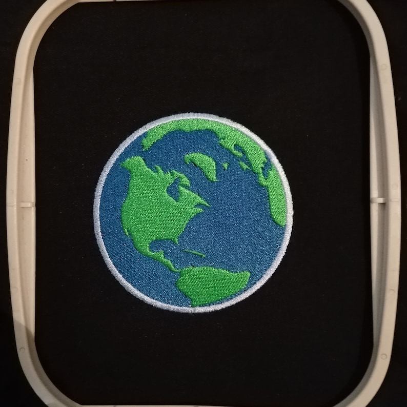 May include: Embroidered patch showing the Earth, with green continents and blue oceans. The circular patch has a white border and is set against a black background within an embroidery hoop. Suitable for customising jackets, bags, or other fabric items.