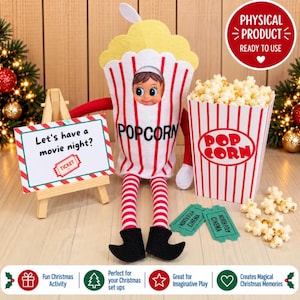 May include: A plush elf doll wearing a popcorn bucket costume with red and white stripes, featuring a cutout for the face. Beside it sits a matching popcorn bucket filled with popcorn, two green cinema tickets, and a sign reading Let's have a movie night?