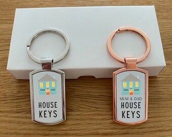 May include: Two house keychains, one silver and one rose gold. Both keychains feature a small house icon and the text "HOUSE KEYS". The rose gold keychain also has the text "MUM & DAD" above the house icon.