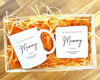 May include: A white mug and a white coaster with the text "The World's most amazing Mummy belongs to Thomas, Joshua & Louisa" printed in black. The mug and coaster are in a basket filled with orange shredded paper.