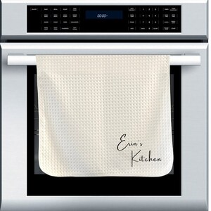 May include: A white waffle weave kitchen towel hanging on a silver oven door handle. The towel has the text "Erin's Kitchen" written in black cursive.