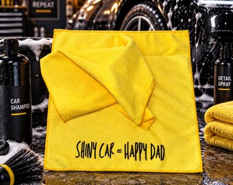 May include: A bright yellow microfiber cleaning cloth featuring the black text SHINY CAR = HAPPY DAD printed on the front. The square cloth is folded over itself and is positioned among various car detailing supplies and cleaning equipment.