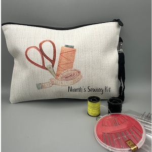 May include: A white canvas zippered pouch with a black zipper and a black strap. The pouch has a printed design of scissors, thread, and a measuring tape. The text "Niamh's Sewing Kit" is printed on the pouch. The pouch is accompanied by a yellow spool of thread, a black spool of thread, and a clear plastic container with red needles.