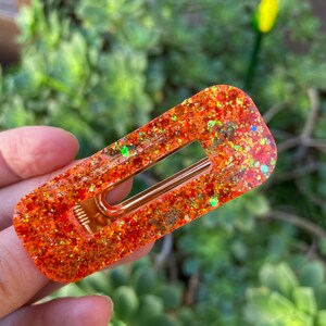 May include: Orange glitter hair clip with a gold metal bar. The clip is rectangular and has a glossy finish.