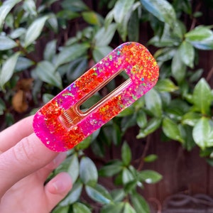 Puede incluir: Una pinza para el pelo de color rosa y naranja con purpurina y un resorte de metal dorado. La pinza tiene forma de rectángulo con bordes redondeados.