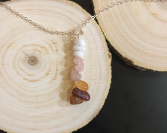 May include: A silver chain necklace with a pendant made of stacked, natural gemstones. The stones are white, pink, orange, and brown. The necklace is displayed on a wooden surface.