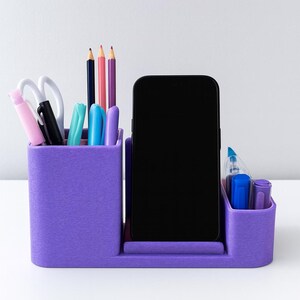 May include: A purple desk tidy with sections for pens, pencils, scissors, and a smartphone. The tidy contains various coloured pens, pencils, and scissors. The smartphone is black. The tidy sits on a white surface.