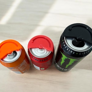 May include: Three beverage cans with coloured lids. An orange can, a red Coke Zero can, and a black Monster Energy can are visible. The lids are designed to cover the opening of the cans. The cans are on a light-coloured surface.