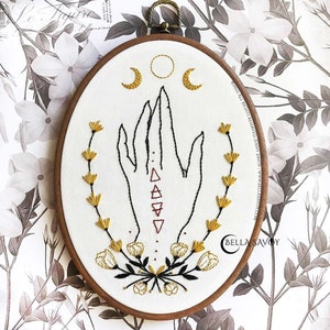 May include: Embroidered hoop art featuring a hand with a red geometric design, surrounded by yellow floral sprigs and a crescent moon design above. The text "Bella Savoy" is in the lower right corner.