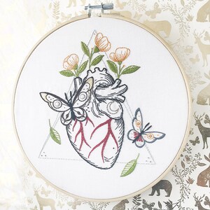 May include: Embroidered hoop art featuring a detailed anatomical heart with red veins, surrounded by delicate green leaves and orange flowers. Two butterflies, one black and white and one with red and blue wings, are also embroidered on the white fabric.