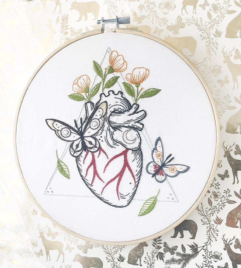 May include: Embroidered hoop art featuring a detailed anatomical heart with red veins, surrounded by delicate green leaves and orange flowers. Two butterflies, one black and white and one with red and blue wings, are also embroidered on the white fabric.