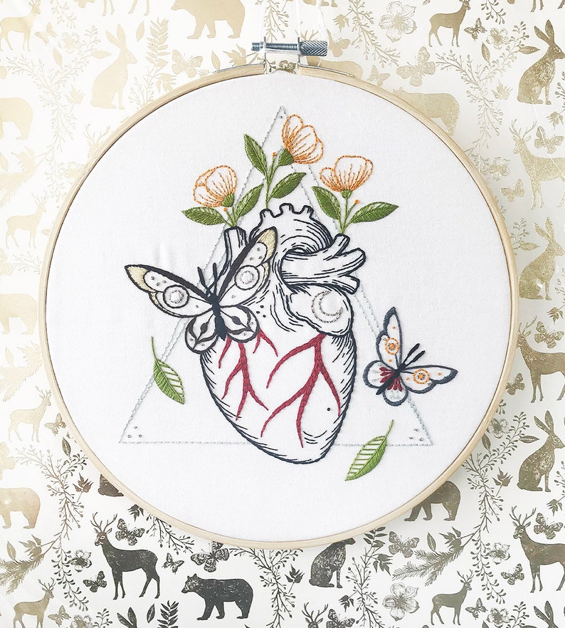 May include: Embroidered hoop art featuring a detailed anatomical heart with red veins, surrounded by delicate flowers and butterflies. The heart is set within a triangle shape.