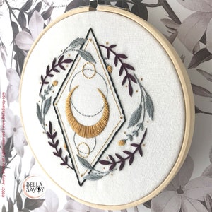May include: Embroidered hoop art featuring a gold crescent moon with geometric shapes and a wreath of burgundy and gray leaves. The hoop is white with the text "BELLA SAVOY" in black.