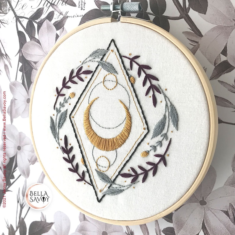 May include: Embroidered hoop art featuring a gold crescent moon with geometric shapes and a wreath of burgundy and gray leaves on a white background.