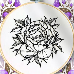 Può includere: Un telaio da ricamo circolare in legno contenente tessuto bianco con il contorno in filo nero di una rosa che presenta un piccolo teschio al centro. Il testo BELLA SAVOY è stampato in nero in alto, con un piccolo simbolo di luna crescente viola.