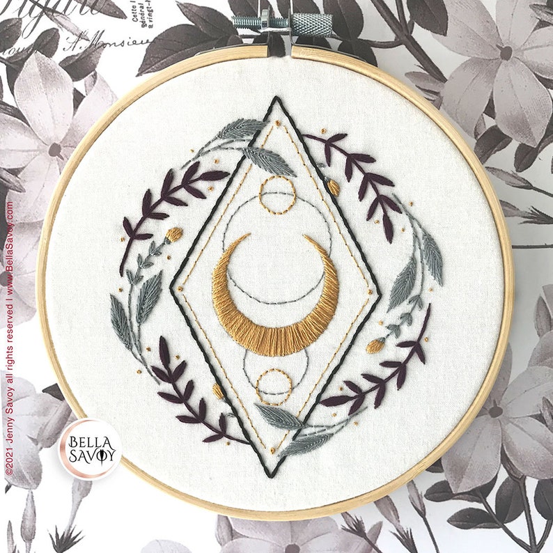 May include: Embroidered hoop art featuring a gold crescent moon with geometric shapes and a wreath of burgundy and gray leaves. The hoop is made of wood and has a white background.