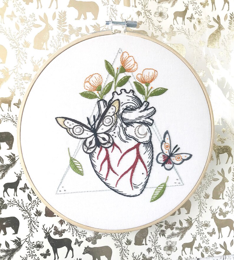 May include: Embroidered hoop art featuring a detailed anatomical heart with red veins, surrounded by delicate orange flowers and two butterflies. The heart is set within a triangle.