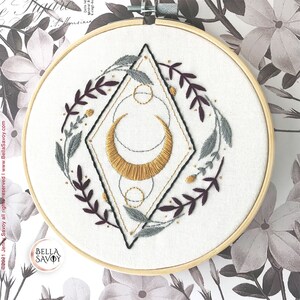 May include: Embroidered hoop art with a gold crescent moon, geometric shapes, and floral sprigs in shades of burgundy and gray. The hoop is made of wood and has a white fabric background.