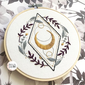 May include: Embroidered hoop art featuring a gold crescent moon inside a geometric diamond shape with a wreath of purple and gray leaves.