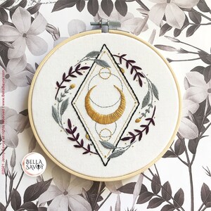 May include: A white embroidery hoop with a gold crescent moon and geometric design surrounded by burgundy and gray leaves. The hoop is on a white background with a floral pattern.