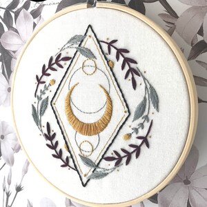 May include: Embroidered hoop art featuring a gold crescent moon with geometric shapes and floral sprigs in shades of burgundy and gray.