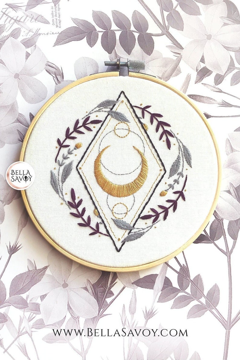May include: Embroidered hoop art featuring a gold crescent moon with geometric shapes and floral sprigs in shades of purple and gray. The hoop is on a white background with a floral pattern.