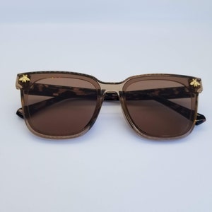 K&ouml;nnte beinhalten: Braune quadratische Sonnenbrille mit Schildpattmuster an den B&uuml;geln und einem goldenen Bienenemblem an jeder Ecke. Die Gl&auml;ser sind einfarbig braun und die Fassungen sind durchscheinend braun.