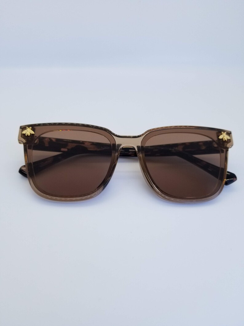 K&ouml;nnte beinhalten: Braune quadratische Sonnenbrille mit Schildpattmuster an den B&uuml;geln und einem goldenen Bienenemblem an jeder Ecke. Die Gl&auml;ser sind einfarbig braun und die Fassungen sind durchscheinend braun.
